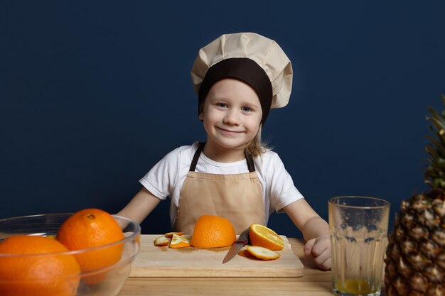 Wesoły mały chłopiec ubrany w fartuch i kapelusz szefa kuchni stojący w nowoczesnej kuchni, gotowanie sałatki owocowej. Portret ładny kaukaski dziecko płci męskiej w mundurze Dokonywanie świeżego soku, cięcia i obierania pomarańczy