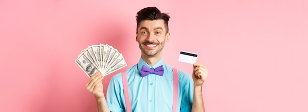 Bezpłatne zdjęcie wesoły mężczyzna pokazujący swoje pieniądze i plastikową kartę kredytową uśmiechający się szczęśliwy do kamery stojącej nad różowymi półdupkami