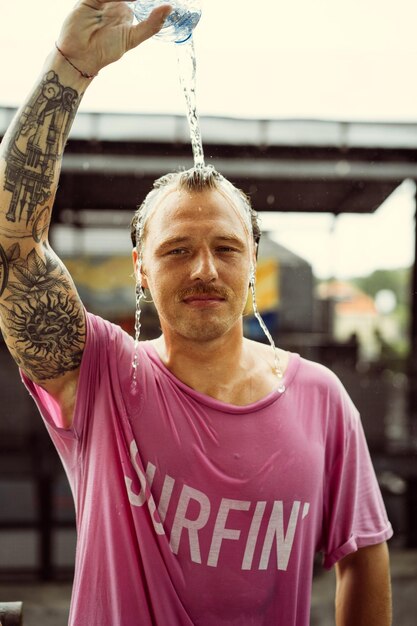 Wesoły mężczyzna w jasnej koszulce z deskorolką w skateparku nalewa sobie wodę z butelki na głowę.