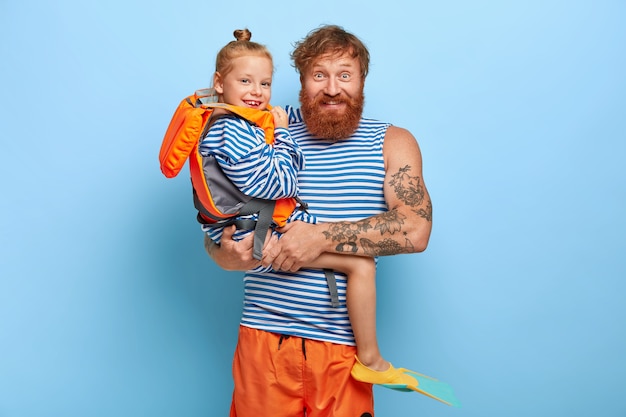 Wesoły młody mężczyzna pozuje z małą rudą dziewczyną, która nosi pomarańczową kamizelkę ratunkową, gumowe płetwy, chętnie spędza letnie wakacje z ojcem, lubi pływać
