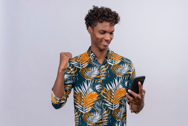 Wesoły młody przystojny ciemnoskóry mężczyzna z kręconymi włosami w koszulce z nadrukiem w liście patrząc na swojego smartfona z zaciśniętą pięścią