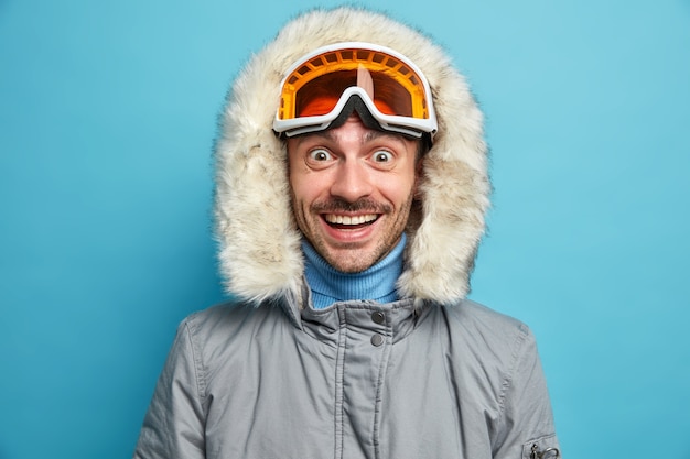 Bezpłatne zdjęcie wesoły nieogolony mężczyzna o radosnym uśmiechu szeroko nosi gogle narciarskie, zimową kurtkę z kapturem lubi ekstremalne sporty zimowe.