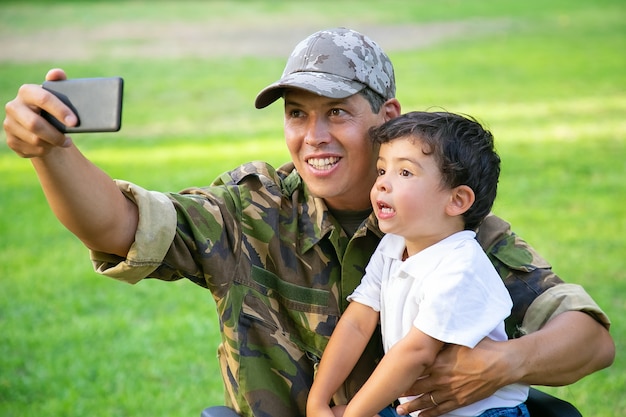 Wesoły niepełnosprawny tata wojskowy i jego synek razem robią selfie w parku. Chłopiec siedzi na kolanach ojców. Weteran wojny lub koncepcji niepełnosprawności