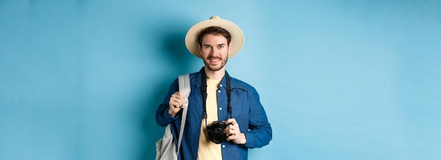 Bezpłatne zdjęcie wesoły przystojny facet jedzie na wakacje w letnim kapeluszu i trzyma plecak z aparatem do zdjęcia
