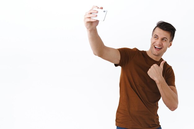 Wesoły przystojny kaukaski mężczyzna w brązowej koszulce biorąc selfie polecam miejsce lub produkt pokaż kciuk jak lub zatwierdź coś trzymającego telefon komórkowy robiąc zdjęcie na smartfonie