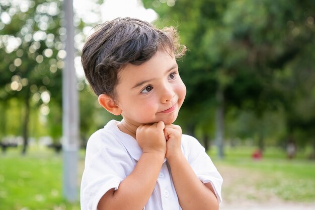 Wesoły słodki chłopiec stojący i pozujący w letnim parku, opierając brodę na rękach, uśmiechając się i odwracając wzrok. Strzał zbliżenie. Koncepcja dzieciństwa