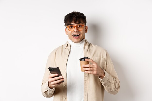 Wesoły uśmiechnięty młody człowiek w okularach picia kawy i trzymając smartfon, patrząc podekscytowany na aparat, stojąc na białym tle.