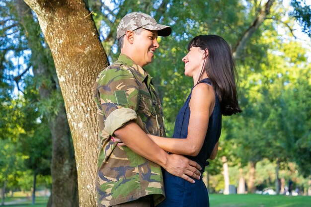 Wesoły wojskowy i jego szczęśliwa żona rozmawiają i przytulają się w parku miejskim. Widok z boku, średni strzał. Koncepcja powrotu do domu lub relacji