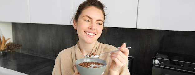 Bezpłatne zdjęcie wesołych poranków, wspaniała młoda kobieta jedząca płatki z mlekiem, stojąca w kuchni z miską śniadaniową