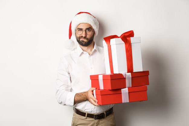 Wesołych Świąt Bożego Narodzenia, koncepcja wakacji. Przemyślany mężczyzna trzyma prezenty świąteczne i patrząc podejrzanie w kamerę, świętuje nowy rok
