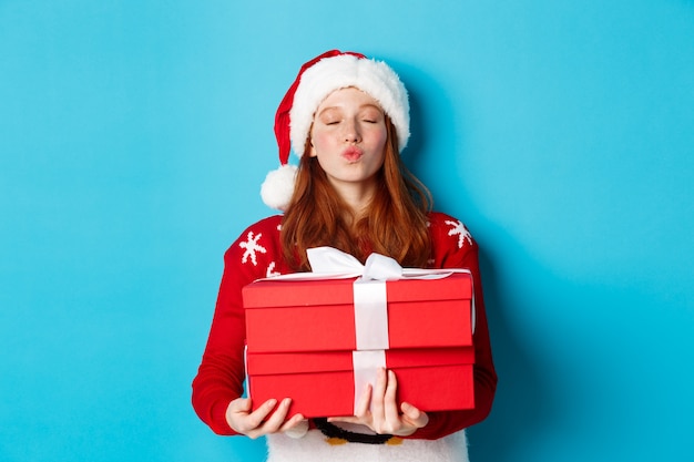 Wesołych Świąt i koncepcji Bożego Narodzenia. Ładna ruda dziewczyna trzyma prezenty i marszczy się usta do pocałunku, nosi czapkę santa i zabawny sweter, niebieskie tło.