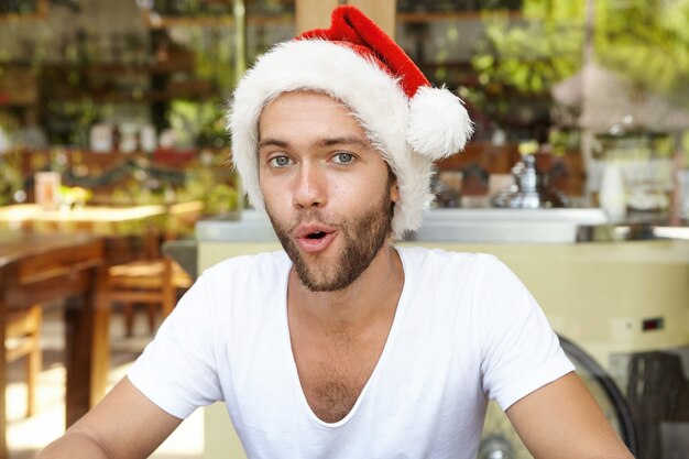 Wesołych Świąt i Szczęśliwego Nowego Roku! Atrakcyjny młody brodaty mężczyzna w czerwonej czapce z białym futrem odpoczywa w pomieszczeniu, udając Świętego Mikołaja, śmiejąc się i otwierając usta, jakby mówił: Ho Ho Ho!