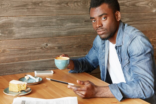 Wewnątrz portret pewny siebie ciemnoskóry mężczyzna ubrany niedbale spędzający weekendowy poranek w kafeterii, siedzący przy drewnianym stole z gadżetami i popijając kawę. Afrykański mężczyzna za pomocą tabletu w kawiarni
