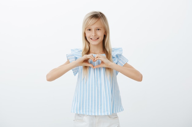 Wewnątrz Portret Uroczej Młodej Dziewczyny O Jasnych Włosach W Niebieskiej Bluzce, Pokazującej Gest Serca Na Piersi I Uśmiechającej Się Ze Szczęścia Na Szarej ścianie