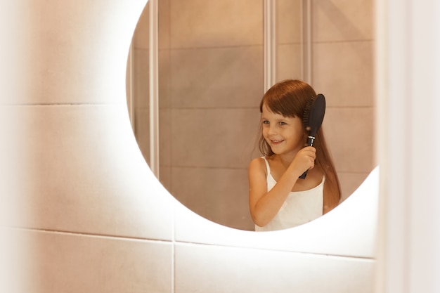 Bezpłatne zdjęcie wewnątrz ujęcie ciemnowłosej dziewczyny czesającej włosy w łazience, robienie porannych zabiegów kosmetycznych przed lustrem, uśmiechnięte szczęśliwie, noszące domowe ubrania w stylu casual.