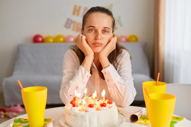 Wewnątrz ujęcie młodej dorosłej pięknej kobiety siedzącej samotnie przy stole, świętującej urodziny tortem i świecami, patrzącej na kamerę, wyrażającej negatywne emocje, czuje się znudzona.