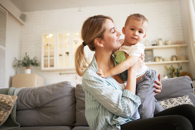 Wewnątrz wizerunek ślicznej młodej kobiety z kucykiem trzymającej mocno swoje urocze dziecko, siedząc z nim na kanapie. Śliczna mama i syn łączą się w salonie, mama patrzy na dziecko z miłością i czułością