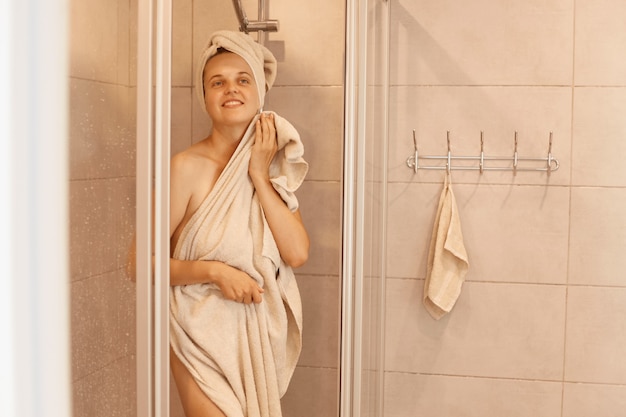 Wewnątrz zdjęcie młodej dorosłej, szczupłej, atrakcyjnej kobiety wychodzącej spod prysznica, stojącej i osuszającej ciało ręcznikiem, patrzącej na kamerę z pozytywnymi emocjami.