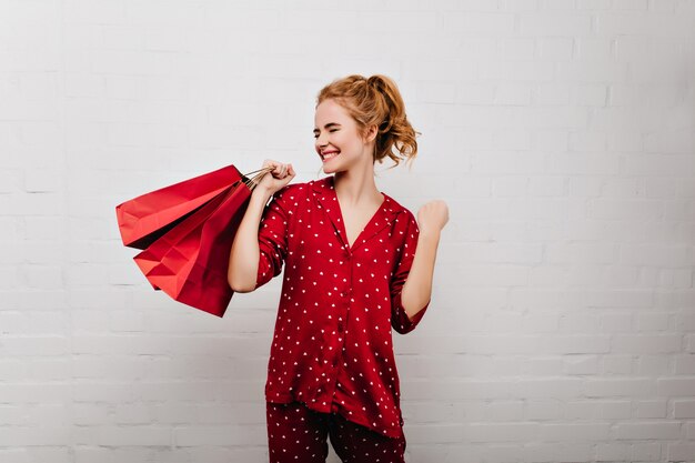 Wewnątrz zdjęcie zadowolonej kobiety rasy kaukaskiej w piżamie z prezentem. czarująca dziewczyna w czerwonym kombinezonie nocnym, śmiejąc się i trzymając papierową torbę.