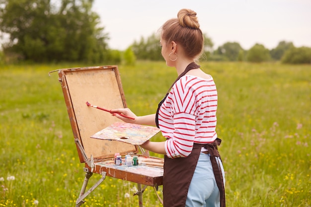 Widok młodej kobiety artysty stojącego przed szkicownikiem z tyłu z pędzlem i paletę kolorów z tyłu
