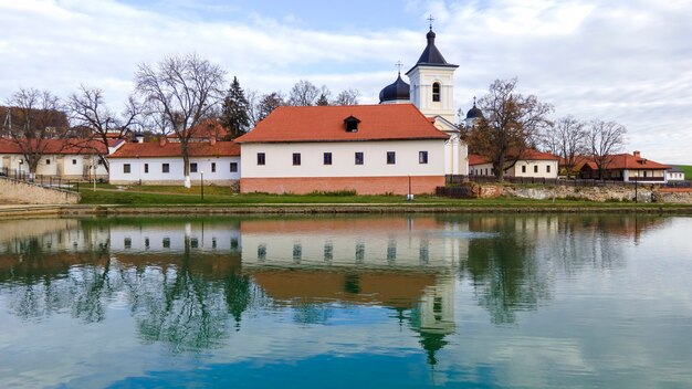 Widok na klasztor Capriana. Kamienny kościół, budynki, nagie drzewa. Na pierwszym planie jezioro, w Mołdawii dobra pogoda