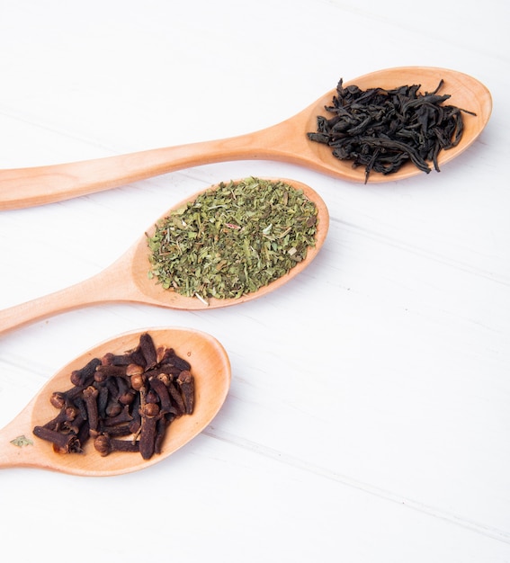 Widok z boku drewniane łyżki z przyprawami i ziołami suszą liście czarnej herbaty, przyprawy goździkowe i suszoną miętę na białym drewnie