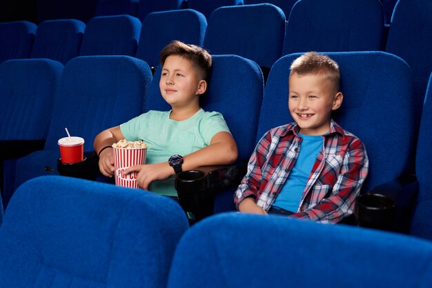 Widok z boku dwóch uśmiechniętych chłopców oglądających razem komiczny film w pustym kinie. Przyjaciele trzymający popcorn i słodką wodę. Dzieci śmieją się i odpoczywają w weekend