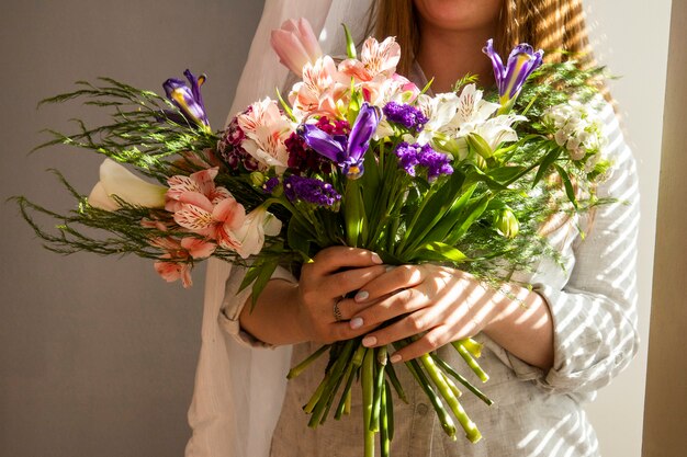 Widok z boku dziewczyny trzymającej bukiet różnych wiosennych kwiatów ciemnofioletowych irysowych kwiatów z alstroemeria, różowych tulipanów, tureckich goździków i fioletowego statice przy lekkim stole