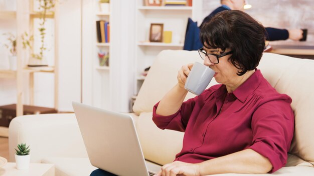 Widok z boku emerytowanej kobiety pisania na laptopie, siedząc na kanapie. Mąż w tle czyta książkę.