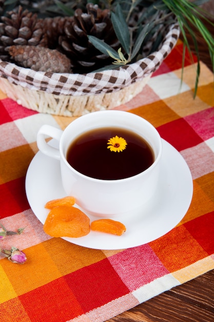Widok z boku filiżankę herbaty z suszonymi morelami i szyszkami sosnowymi w koszu na kraciastym obrusie