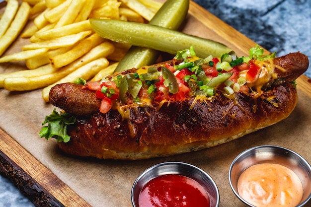 Widok z boku kiełbasa grillowana z hot dogami z zielonymi pomidorami, stopionym serem, papryczką jalapeno w bułce z hot dogami, sosem z frytek i ogórkiem kiszonym na desce