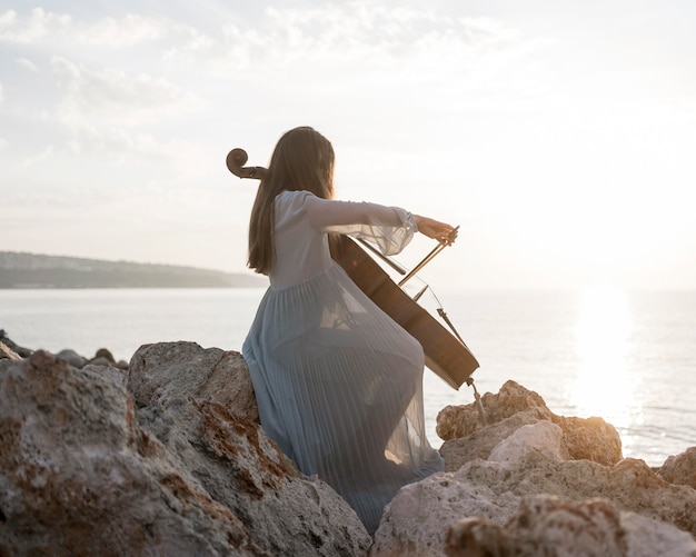 Widok z boku kobiety muzyk gra na wiolonczeli o zachodzie słońca na skałach