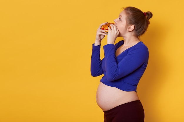 Bezpłatne zdjęcie widok z boku kobiety w ciąży je fast food, ma na sobie niebieską koszulę i legginsy, z wiązką na głowie
