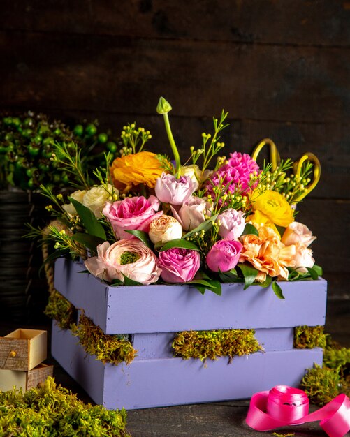 Widok z boku kompozycji różowych i liliowych róż oraz kwiatów ranunculus w drewnianym pudełku