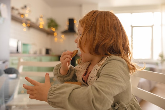 Widok z boku małej jasnoskórej rudej dziewczyny z przyjemnością je ciasteczka, siedząc przy stole w domu Koncepcja żywności i dzieci