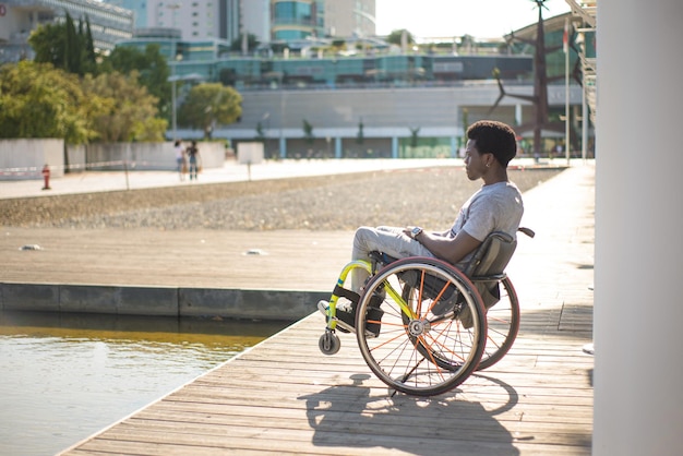 Widok z boku marzycielski mężczyzna na wózku inwalidzkim. African American człowieka w ubranie na nabrzeżu, patrząc na wodę. Miasto w tle. Portret, piękno, koncepcja szczęścia