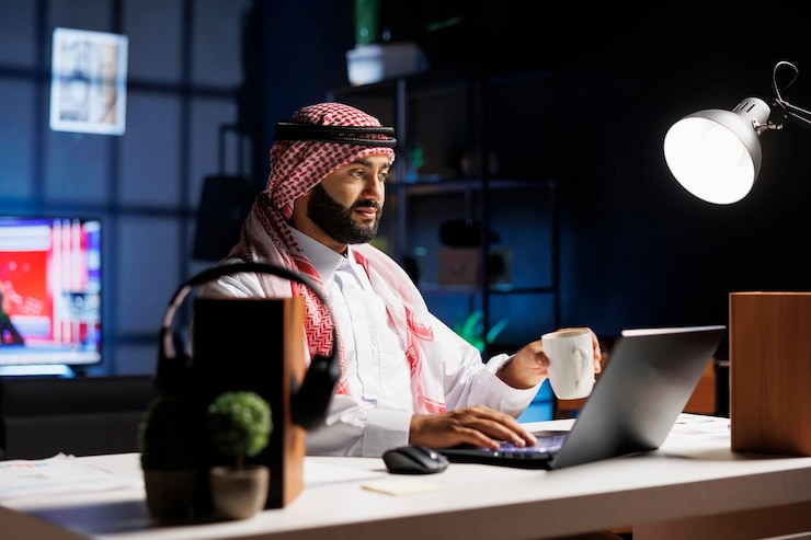 Bezpłatny plik Zdjęcie widok z boku muzułmanina trzymającego filiżankę kawy i surfującego po internecie na swoim cyfrowym laptopie. zdjęcie przedstawiające araba siedzącego przy komputerze podczas picia herbaty.