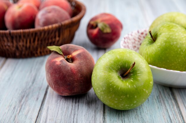 Widok z boku na kolorowe i świeże owoce, takie jak brzoskwinie na wiadrze i jabłko na białej misce na szarej powierzchni drewnianej