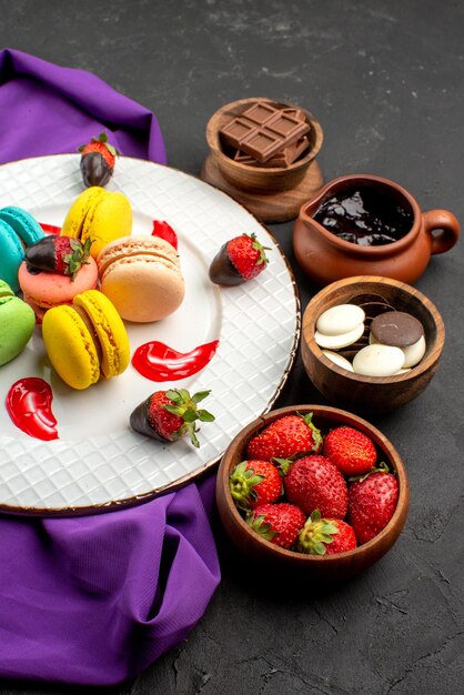 Widok z boku na słodycze Francuskie makaroniki na fioletowym obrusie i cztery miski słodyczy na ciemnej ścianie