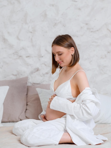 Widok z boku pięknej kobiety ubranej w biały domowy garnitur i biustonosz spodziewa się dziecka, dotykając dużego brzucha i czując ruch siedząc na kolanach na łóżku w mieszkaniu