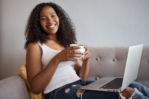 Widok z boku pięknej, uroczej młodej afrykańskiej kobiety po dwudziestce odpoczywającej w domu po studiach, leżącej na kanapie z przenośnym komputerem na kolanach, surfującej po Internecie, zakupów online, uśmiechnięta