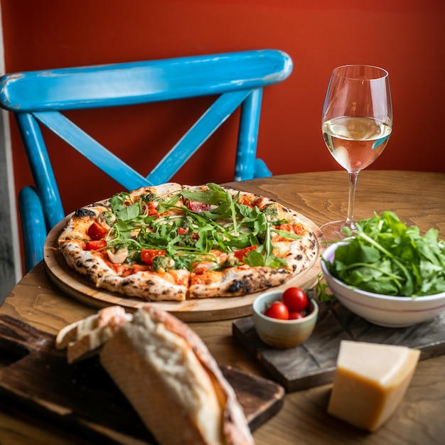 Widok z boku pizzy na drewnianej desce kieliszek białego wina kromka chleba i ser zielony i pomidory w małej białej misce
