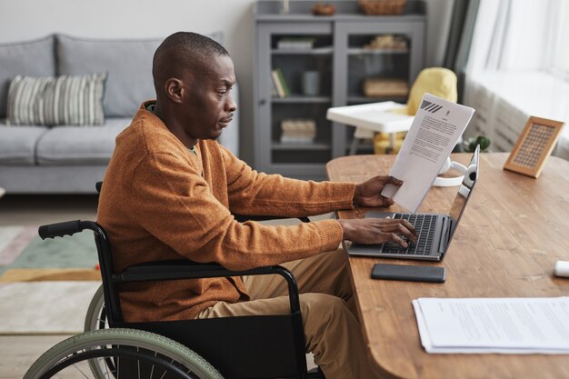 Widok z boku portret afroamerykańskiego mężczyzny korzystającego z wózka inwalidzkiego pracującego w domu w minimalnym szarym wnętrzu, kopia przestrzeń
