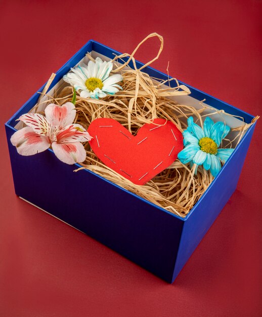 Widok z boku prezentowego pudełka wypełnionego słomą, czerwonym sercem wykonanym z papieru oraz kwiatami stokrotki i alstroemeria na czerwonym stole