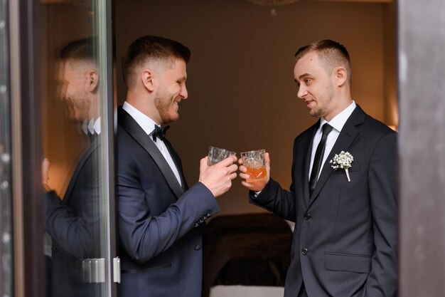 Widok z boku przystojnych mężczyzn w eleganckich garniturach brzęczących okularami świętujących dzień ślubu na wieczorze kawalerskim Stojący w przytulnym mieszkaniu i uśmiechnięty Przyjaciel pana młodego