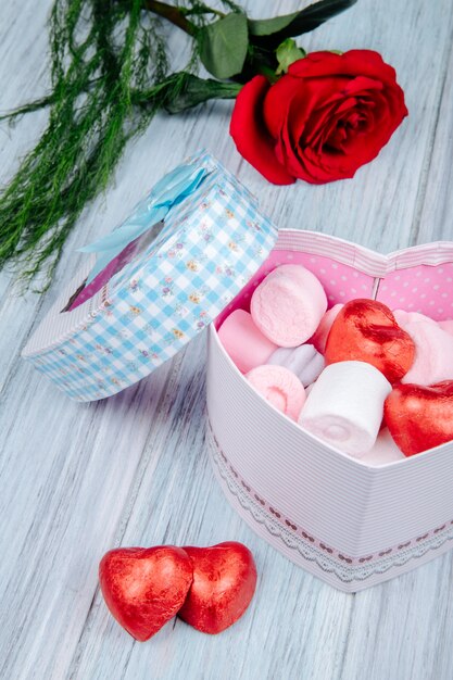 Widok z boku pudełka w kształcie serca wypełnionego różowymi piankami marshmallow i czekoladowymi opakowanymi w czerwoną folię i czerwony kwiat róży na szarym drewnianym stole