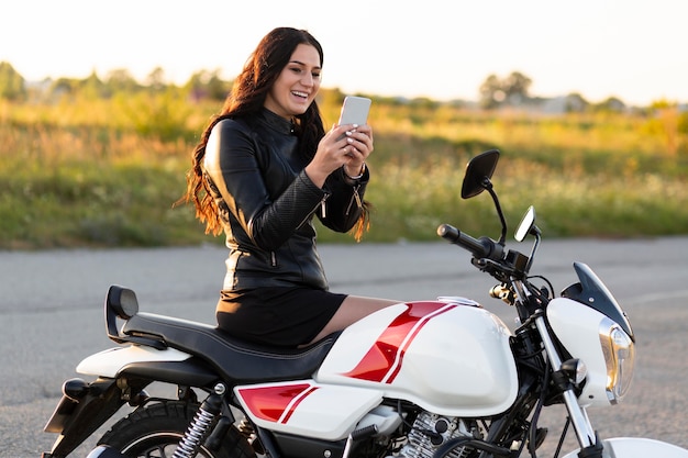 Widok z boku uśmiechniętej kobiety patrząc na smartfona siedząc na swoim motocyklu