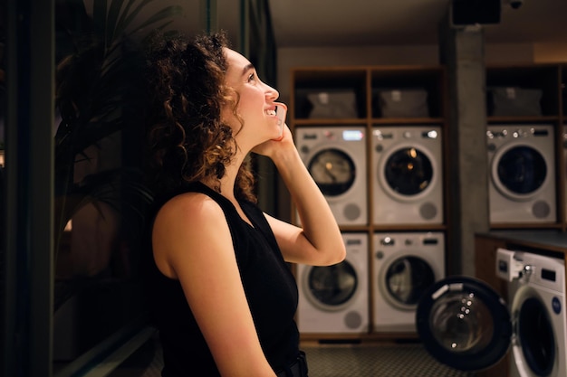 Widok z boku wesołej dziewczyny rozmawiającej na smartfonie czekającej na pranie w nowoczesnej pralni samoobsługowej w nocy