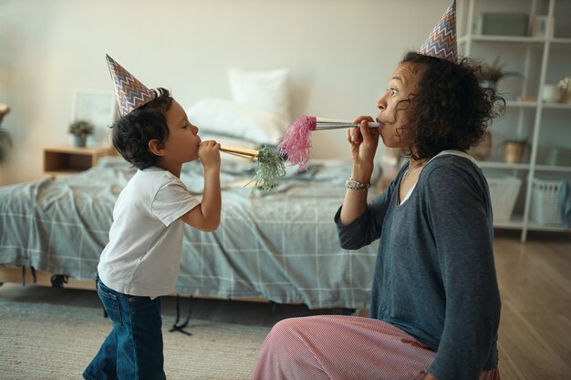 Widok z boku wesołej młodej samicy rasy mieszanej i jej uroczego małego synka w stożkowych czapkach, dmuchających w gwizdek podczas świętowania urodzin samotnie w domu podczas kwarantanny