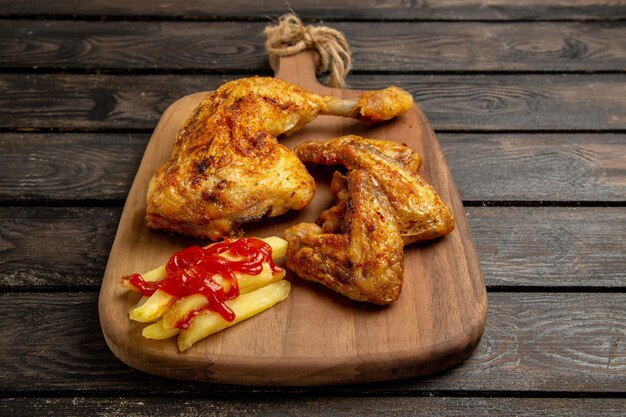 Widok z boku z bliska kurczak i przyprawy skrzydełka i udko z kurczaka z frytkami i keczupem na desce do krojenia na środku ciemnego stołu